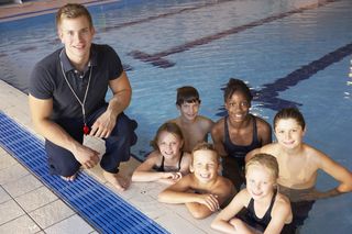 Farbfoto: Ein junger Mann hockt am Rand eines Schwimmbeckens. Neben ihm im Wasser stehen sechs Kinder und blicken in die Kamera.