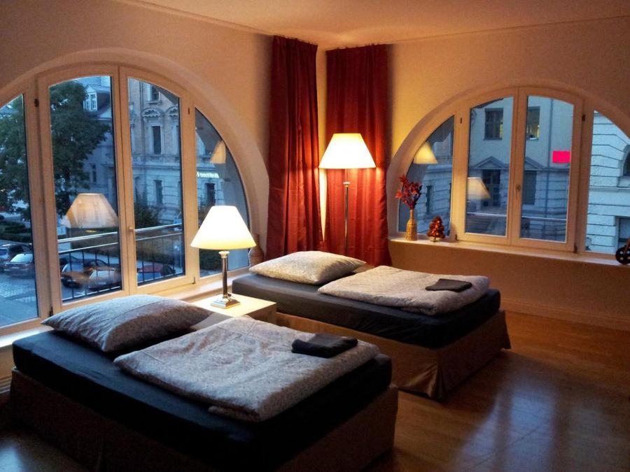 zur Vergrößerungsansicht des Bildes: Farbfoto: Innenansicht eines Wohnraums mit zwei Betten, bodentiefen Fenstern und einer Stehlampe