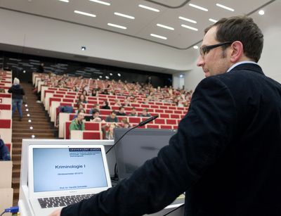 Foto: ein Mann steht am Rednerpult im voll bestetztem Hörsaal und spricht zu den Studierenden während er auch auf einen Laptop blickt.