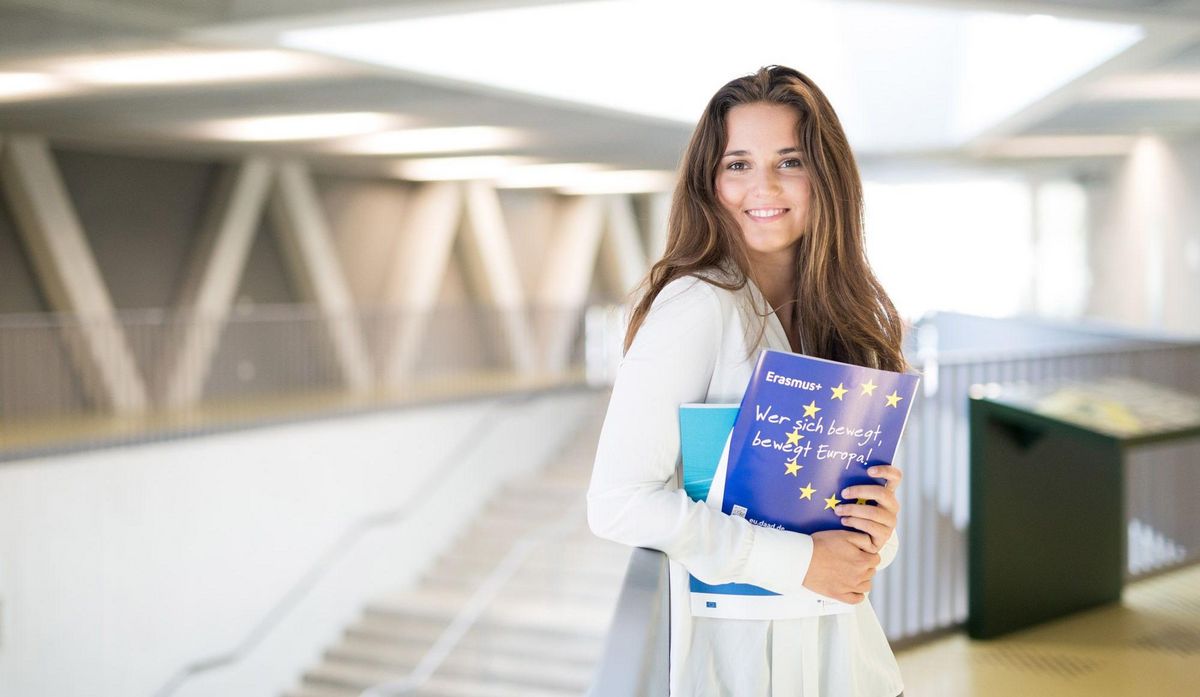 enlarge the image: Junge Frau mit Erasmus Prospekten steht in der Uni