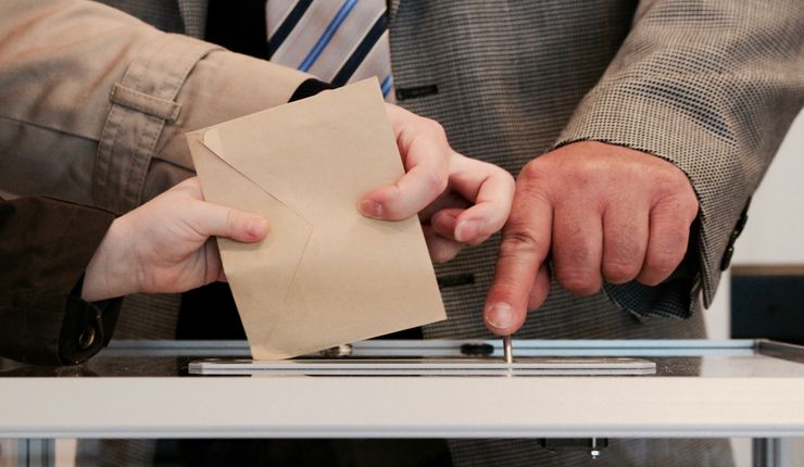 Two hands post an envelope into a ballot box.